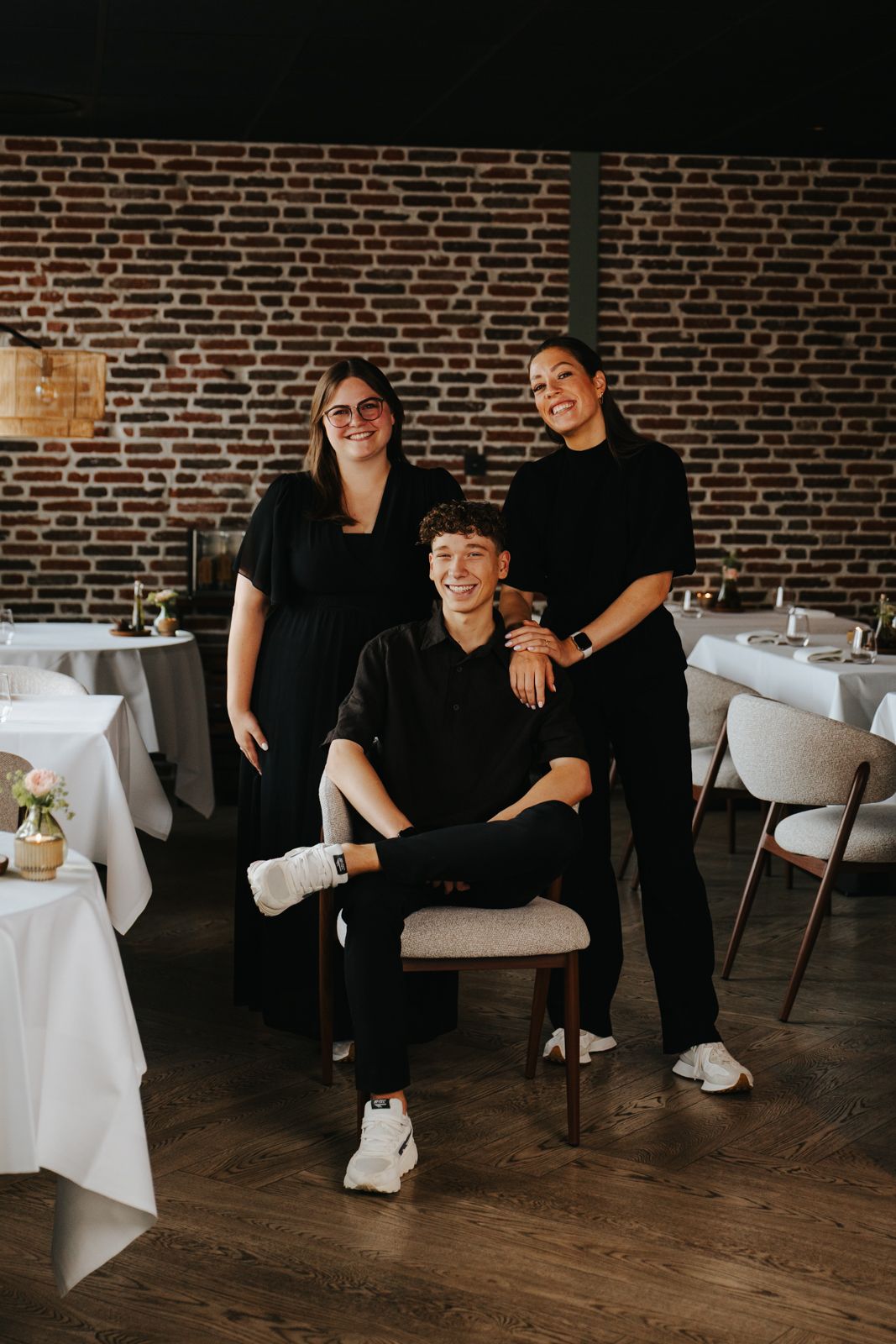 Three people dressed in black posing in an elegant restaurant with white tablecloths and exposed brick walls.