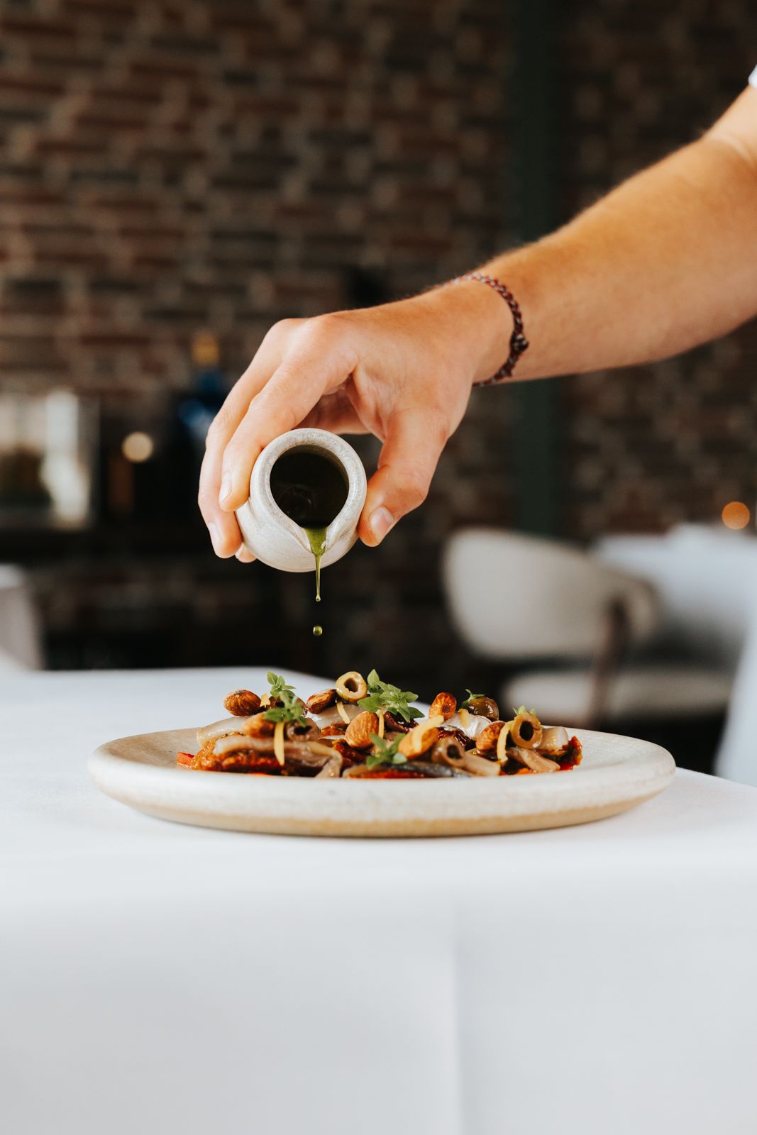 A hand pouring dark sauce from a small white pitcher onto a gourmet dish on a white tablecloth in a restaurant.