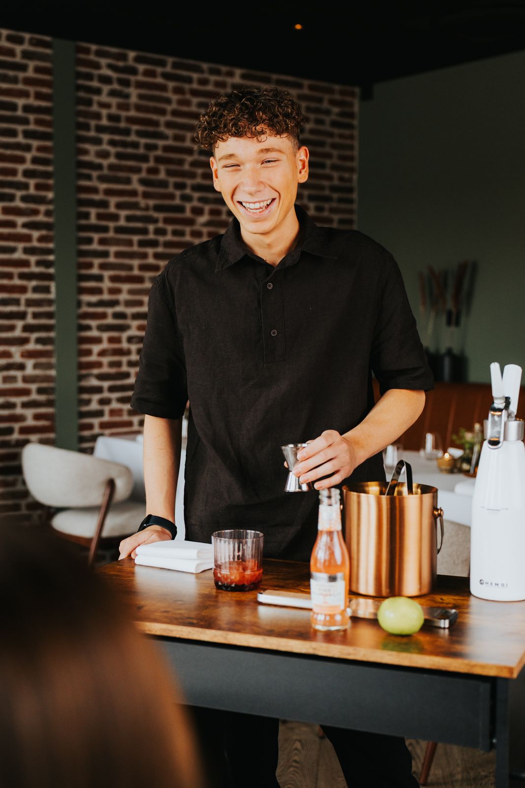 Smiling young bartender in black shirt preparing cocktails at a wooden bar counter with copper shaker and bottles.
