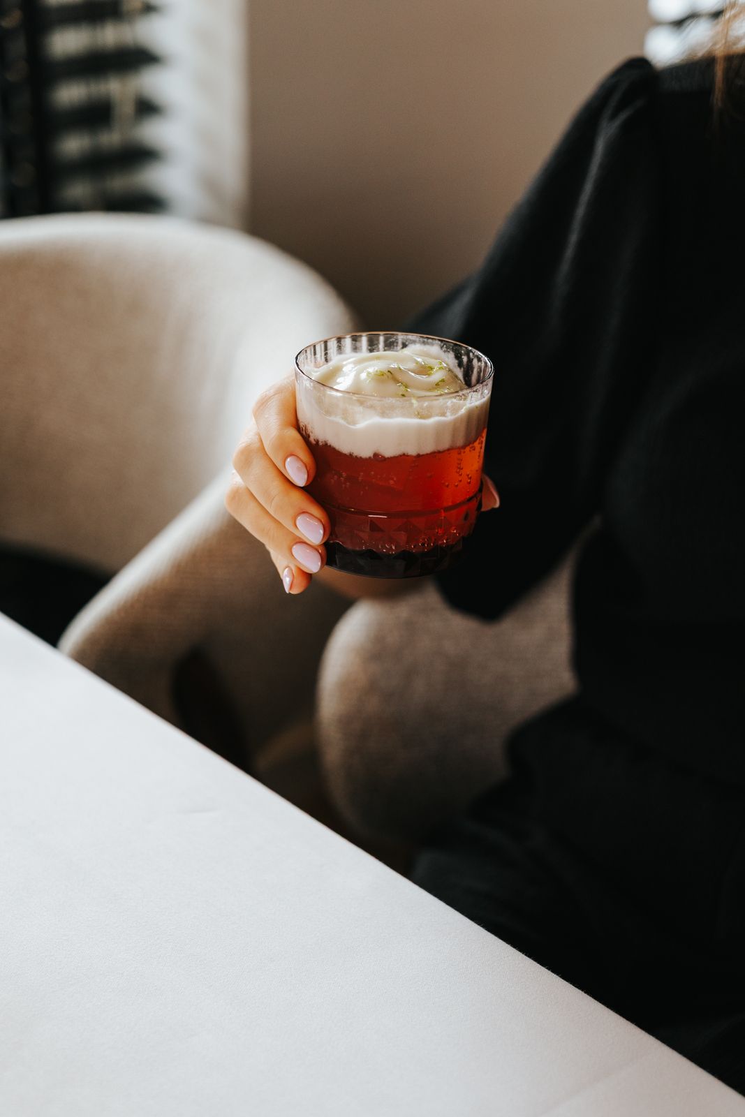 Woman in black sweater holding a red cocktail with whipped cream and lime garnish while seated in a beige chair.