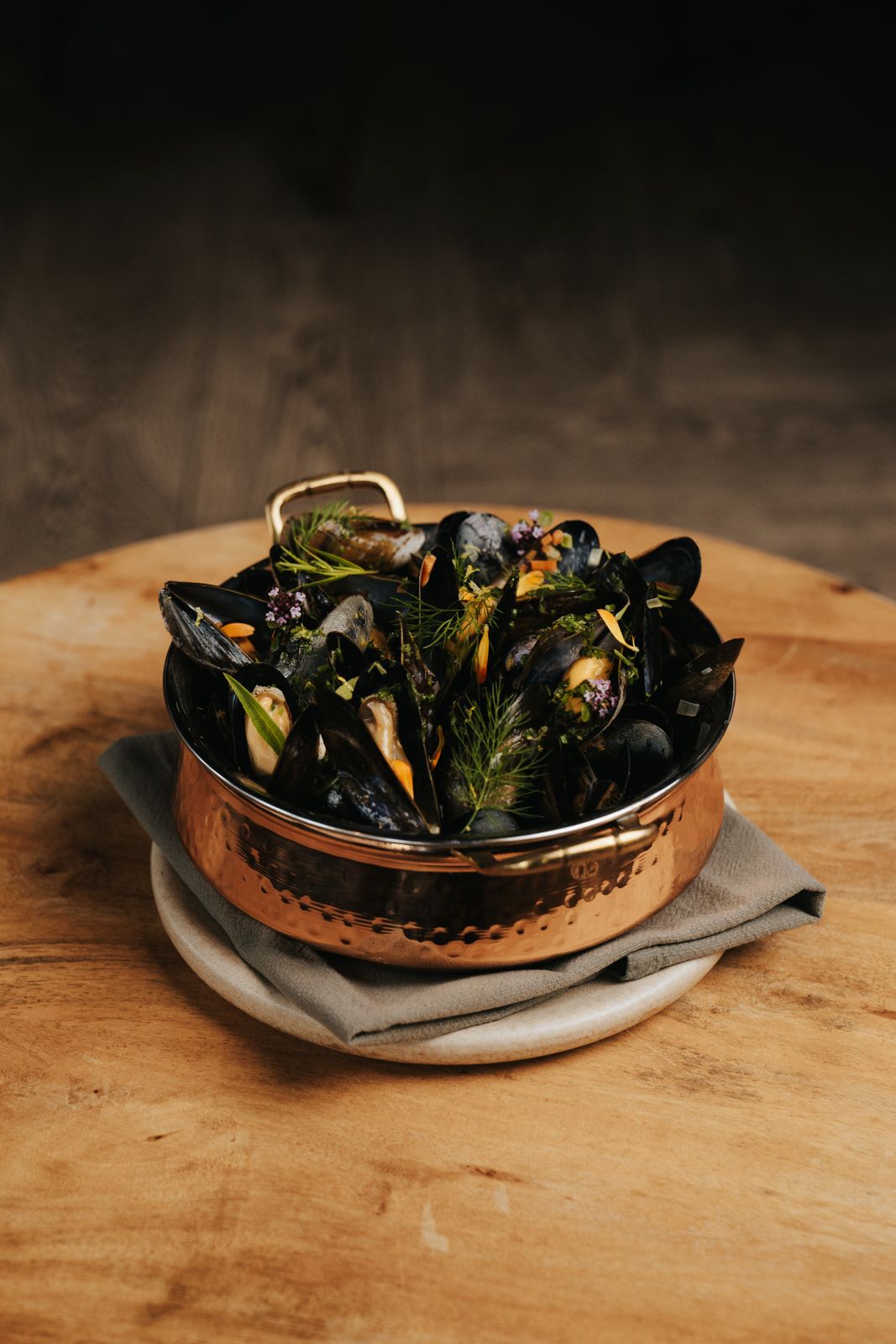 A copper bowl filled with steamed mussels garnished with fresh herbs, served on a linen napkin on a wooden board.
