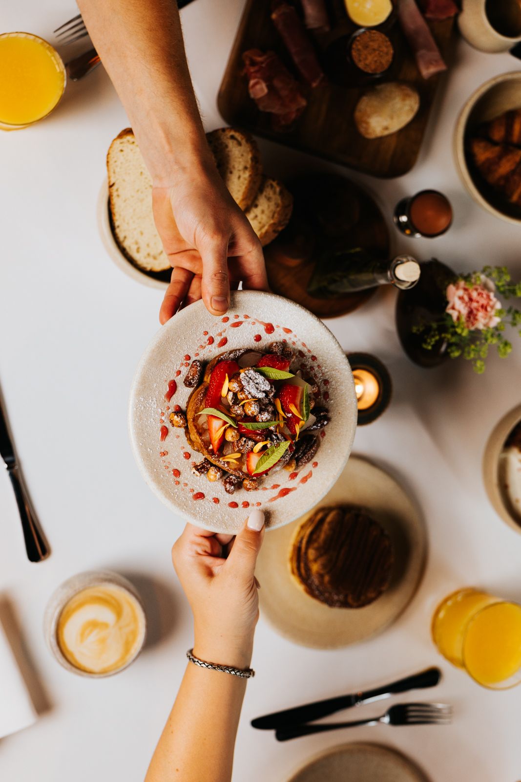 Two hands passing a colorful gourmet dish over a brunch table with orange juice, bread, and candles.