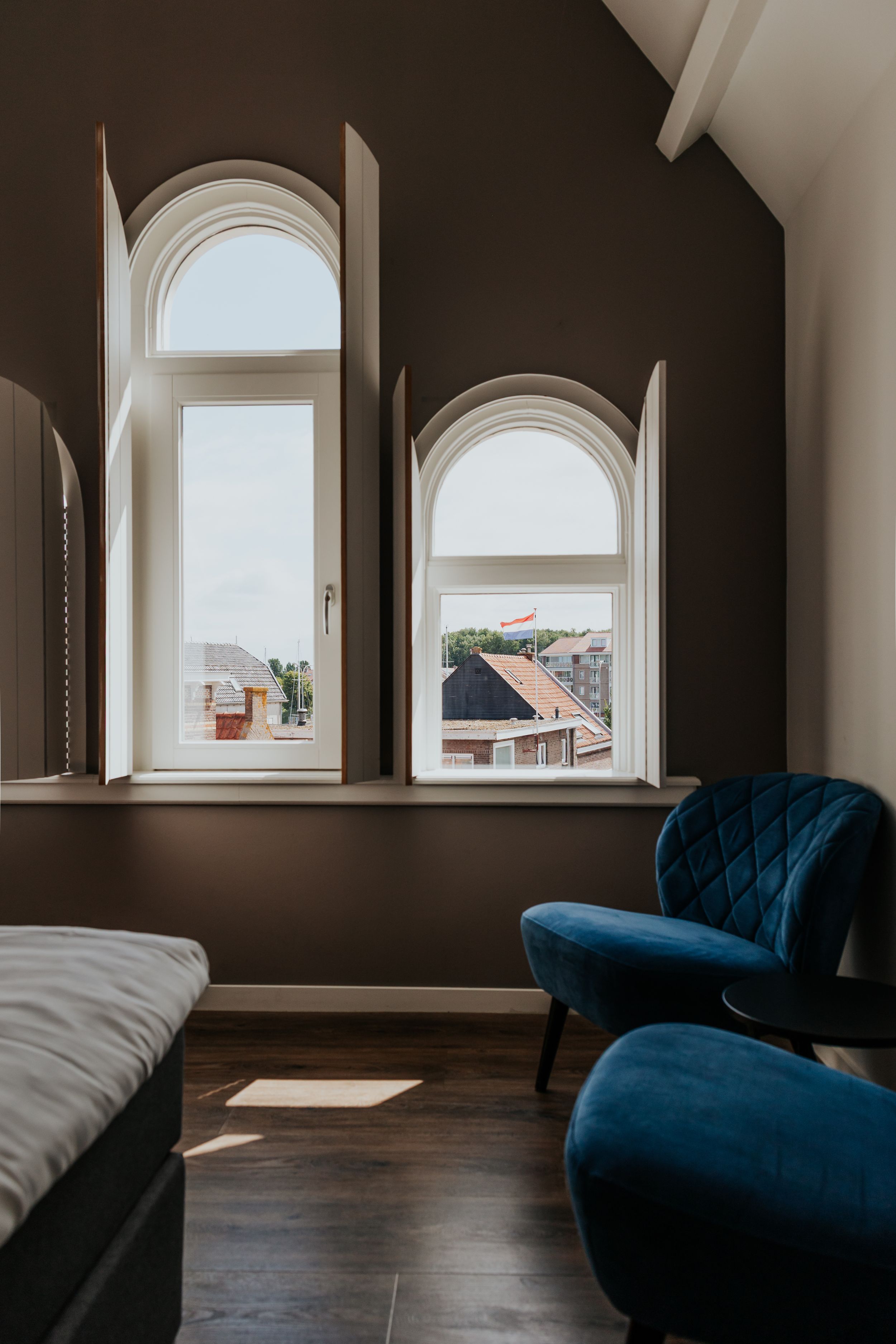 Modern bedroom interior with arched windows, dark walls, and blue velvet accent chair in minimalist design.