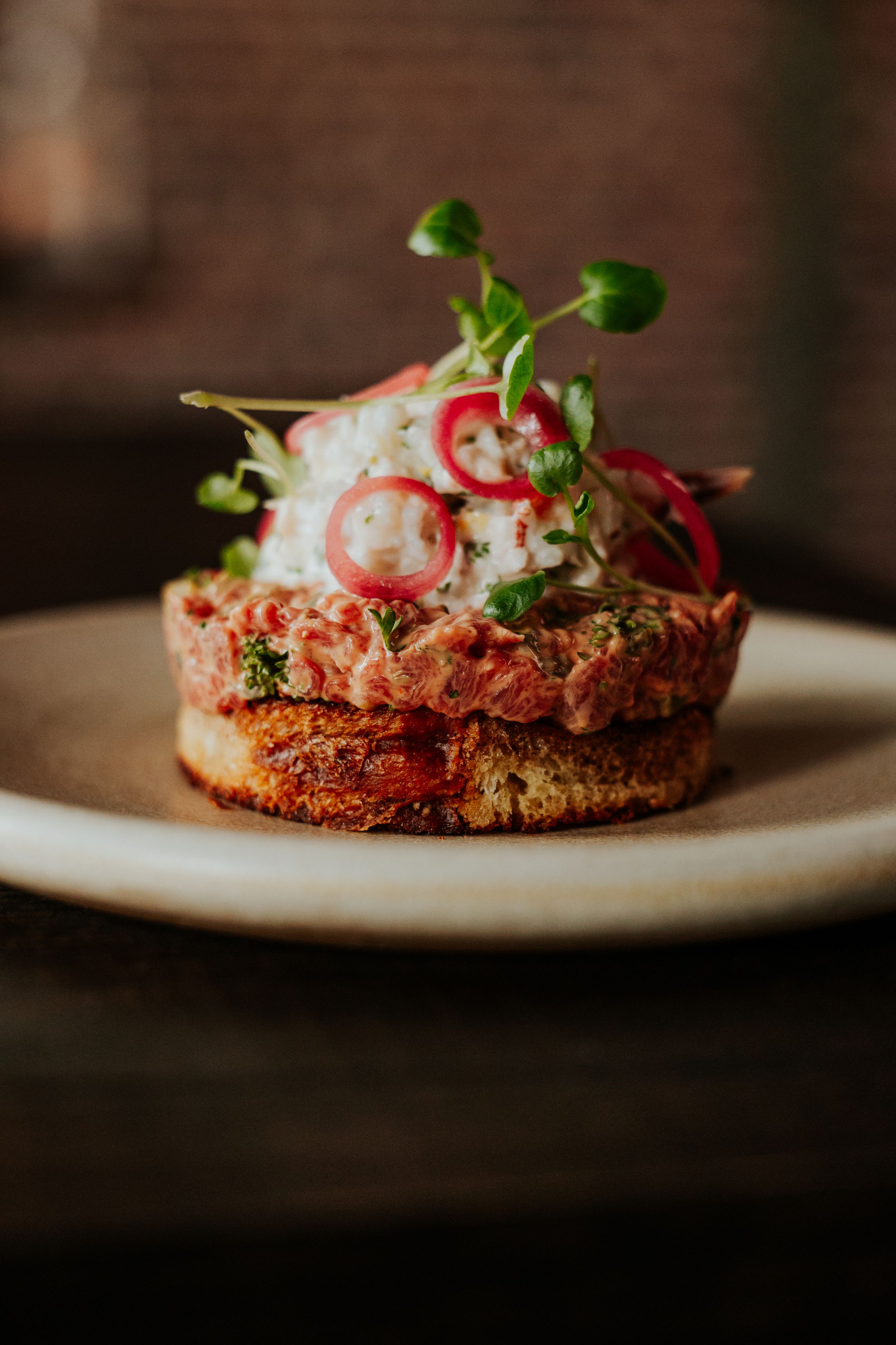 Gourmet appetizer plated on white dish with microgreens and red garnish in rustic setting.