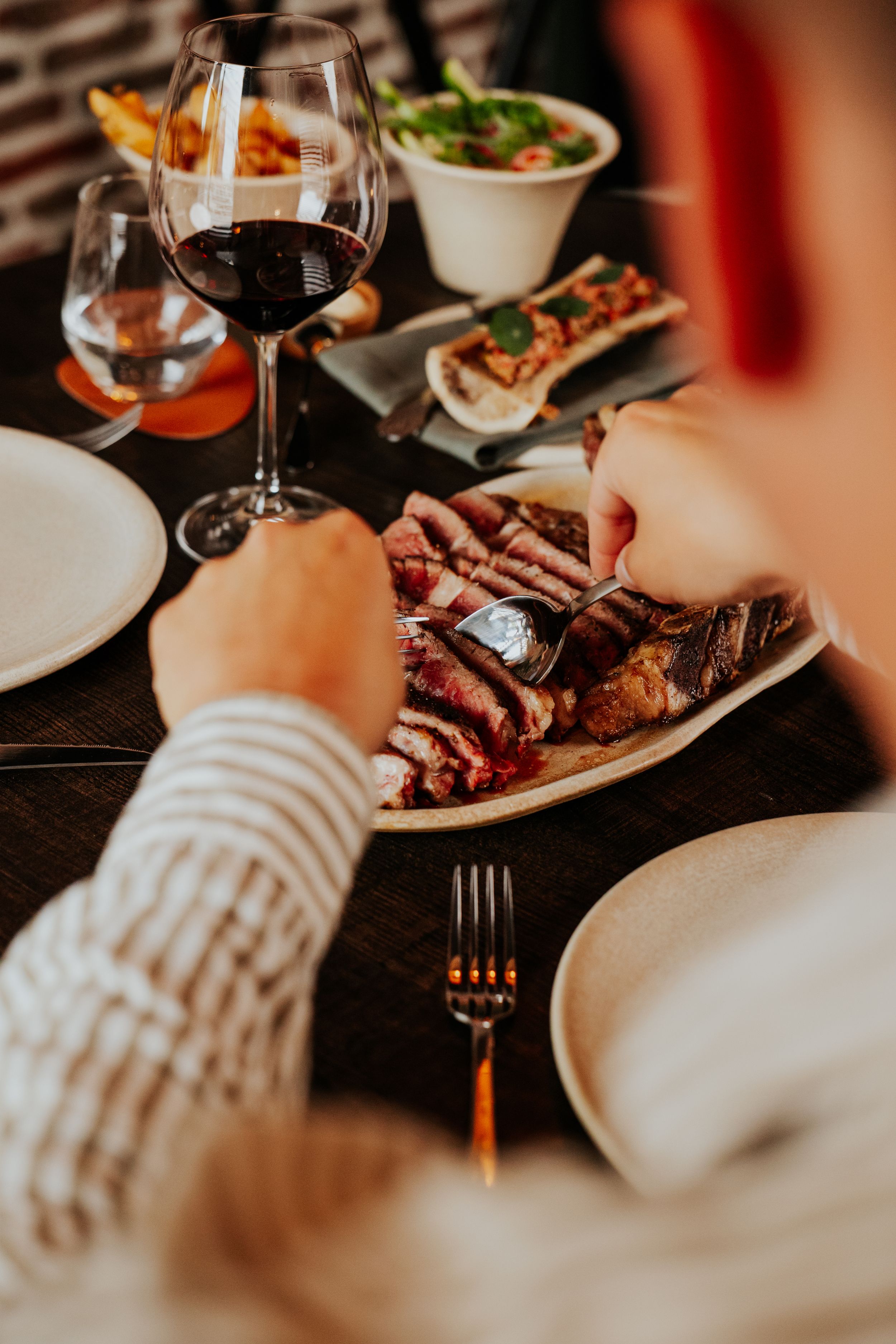 Person cutting into perfectly cooked steak with wine glass on dark table setting.