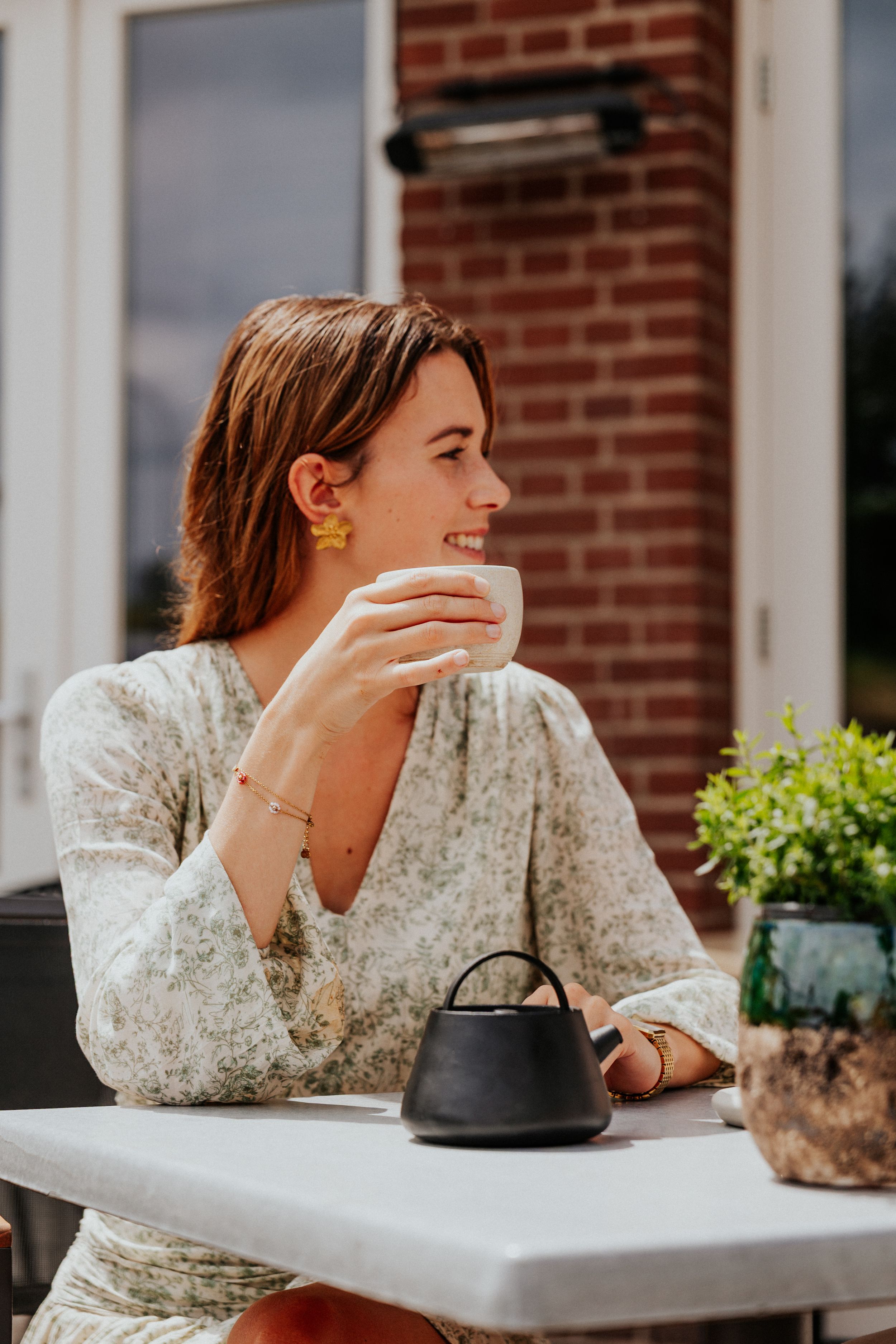 Woman in white floral dress smiling while sitting at outdoor cafe table with teapot.