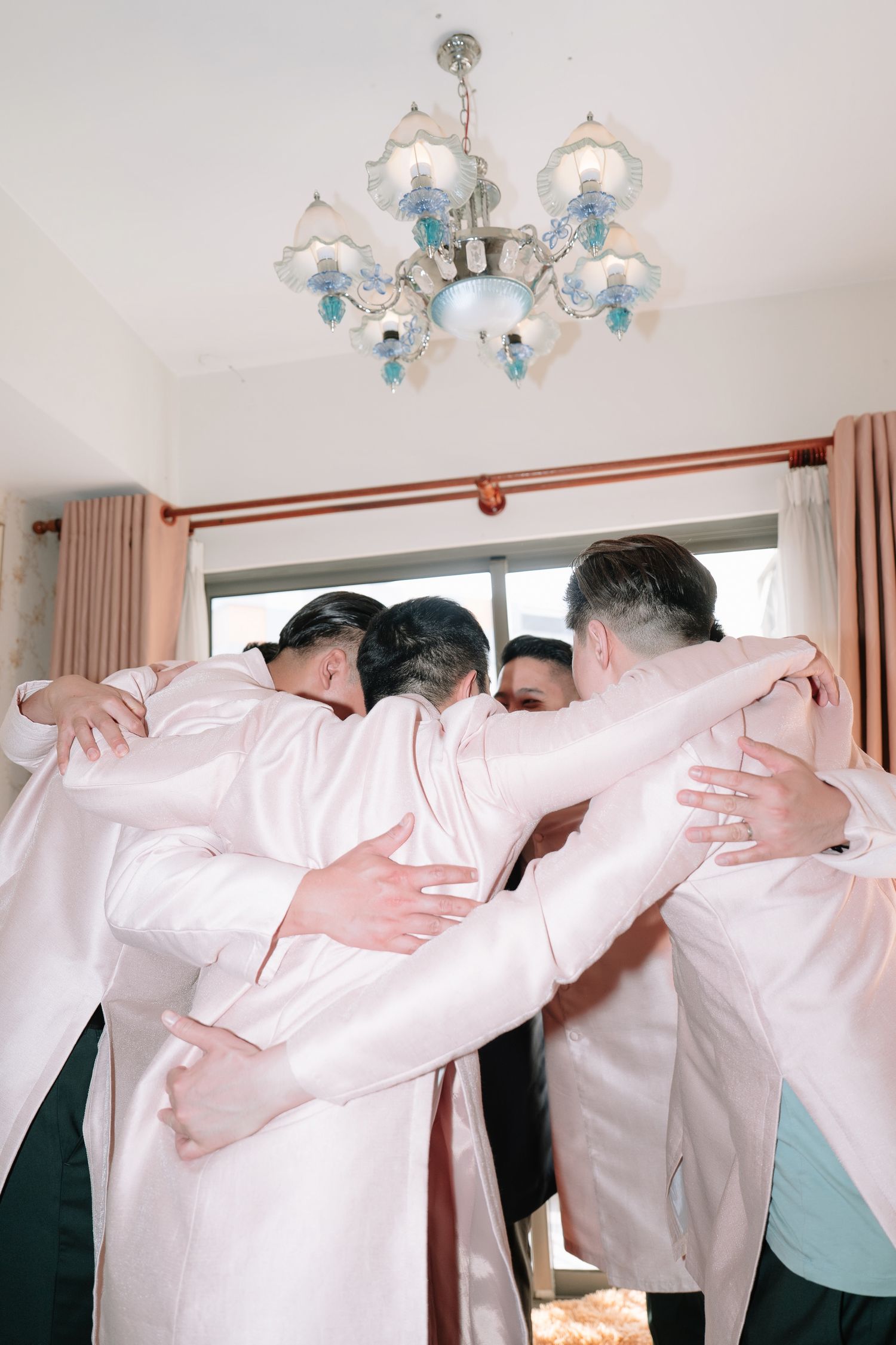 Groomsmen in pink ao dai sharing a group hug under an elegant chandelier before a wedding.