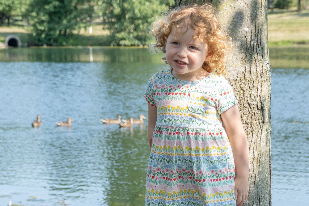 A toddler plays near a lake with ducks swimming in the background on a sunny summer day.