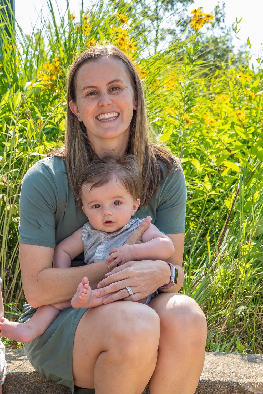 A mother and baby sit together among yellow wildflowers on a sunny summer day outdoors.
