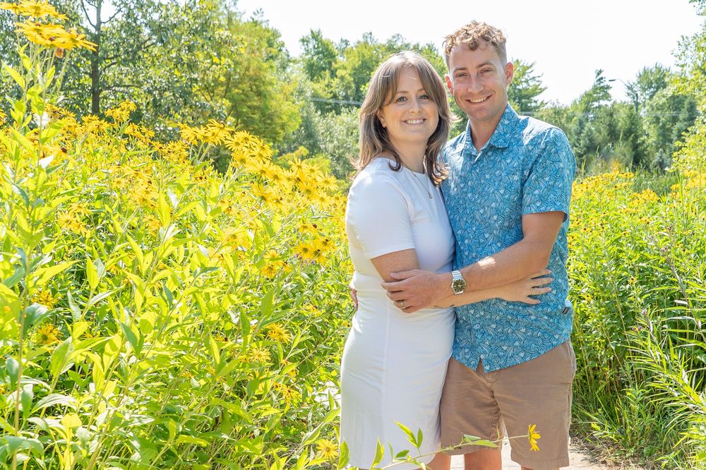 A couple poses together in front of vibrant yellow wildflowers on a sunny afternoon in a natural setting.