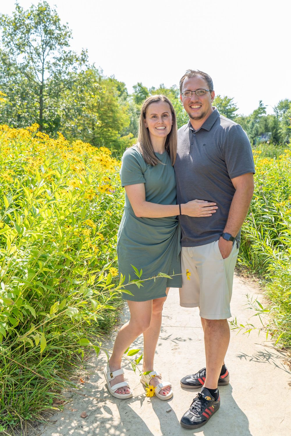 Couple stands together on walking path surrounded by vibrant yellow wildflowers on a sunny summer day.