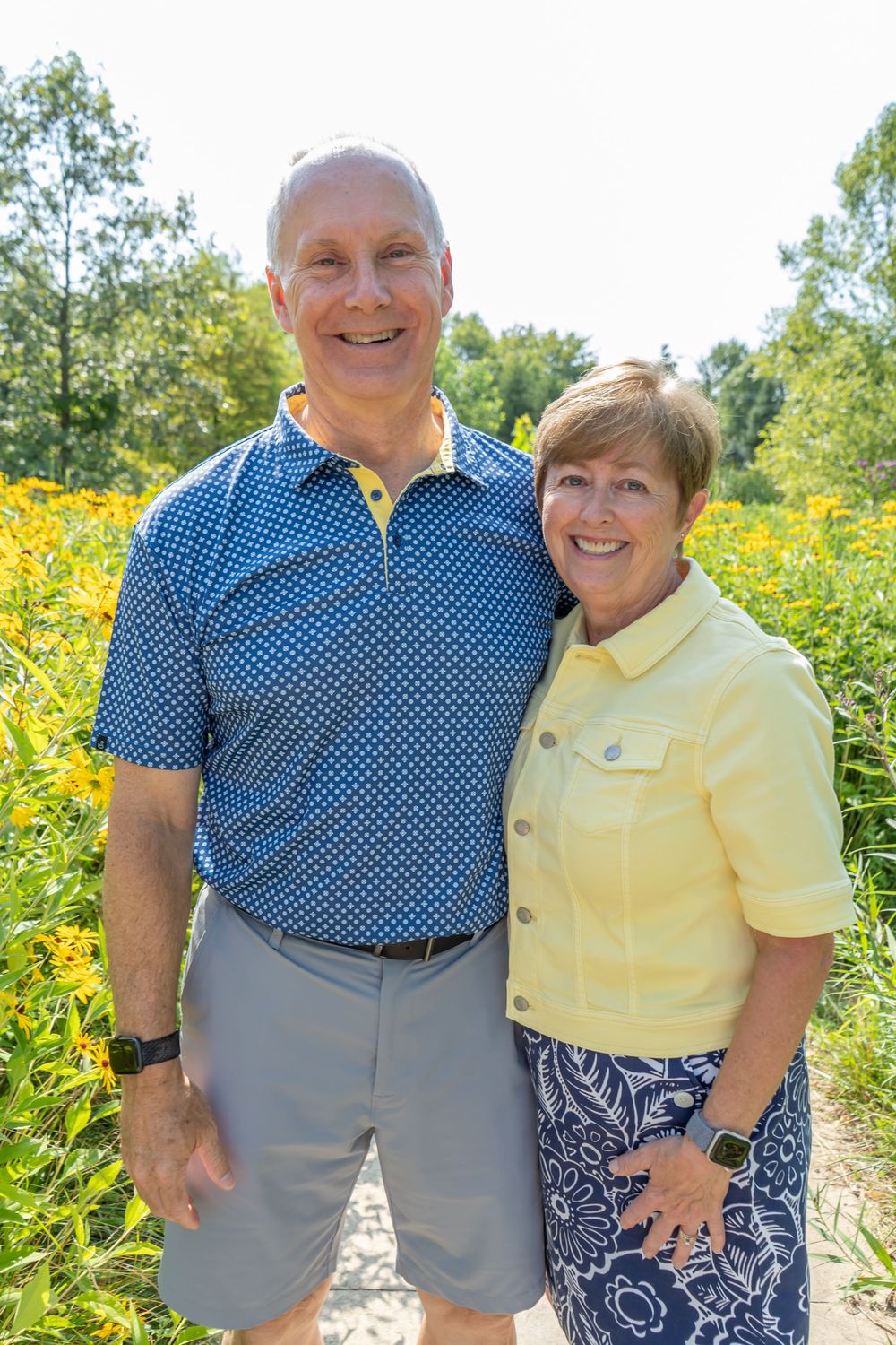 Two people wearing blue polka dot and yellow outfits pose outdoors with yellow wildflowers in the background.