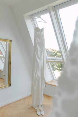 Long white wedding dress hanging by a sunlit window in an attic room.