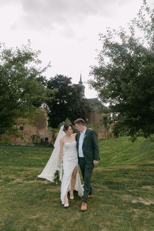 A couple in wedding attire walks together on a grassy hillside with trees and buildings in the background.