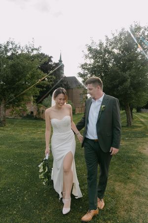 A couple shares a playful moment on their wedding day while walking through a grassy field.
