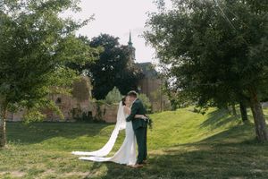 Wedding couple strolls across a scenic grassy hillside with trees and architecture visible in the distance.