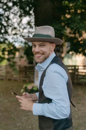 Person in hat and dress shirt smiling while looking at phone outdoors.