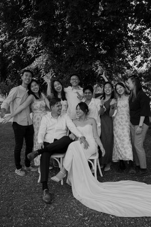 Wedding party poses for fun group photo on lawn with dress train visible.
