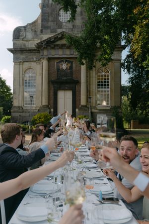 Long outdoor wedding reception table with guests toasting against historic building backdrop.