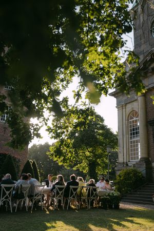 Outdoor dining setup at sunset with guests seated under tree canopy near a historic building.