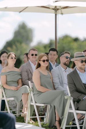 Wedding guests seated in white chairs under a sunshade wearing coordinated gray attire.