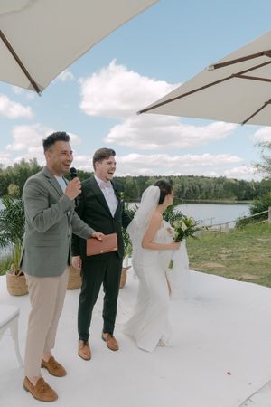 Outdoor wedding ceremony by a lake with white umbrellas and guests gathered on a sunny day.