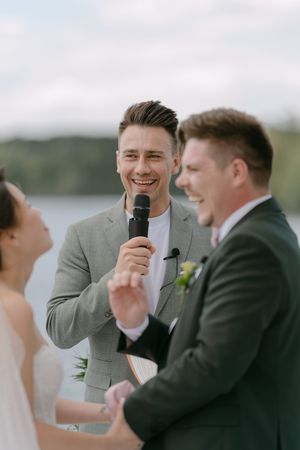 Wedding officiant speaks into microphone during lakeside ceremony while couple shares a joyful moment.