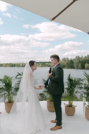 Couple exchanges vows during lakeside ceremony surrounded by potted palms and wicker baskets.
