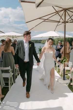 Newlyweds walk hand in hand under white umbrellas after lakeside ceremony.