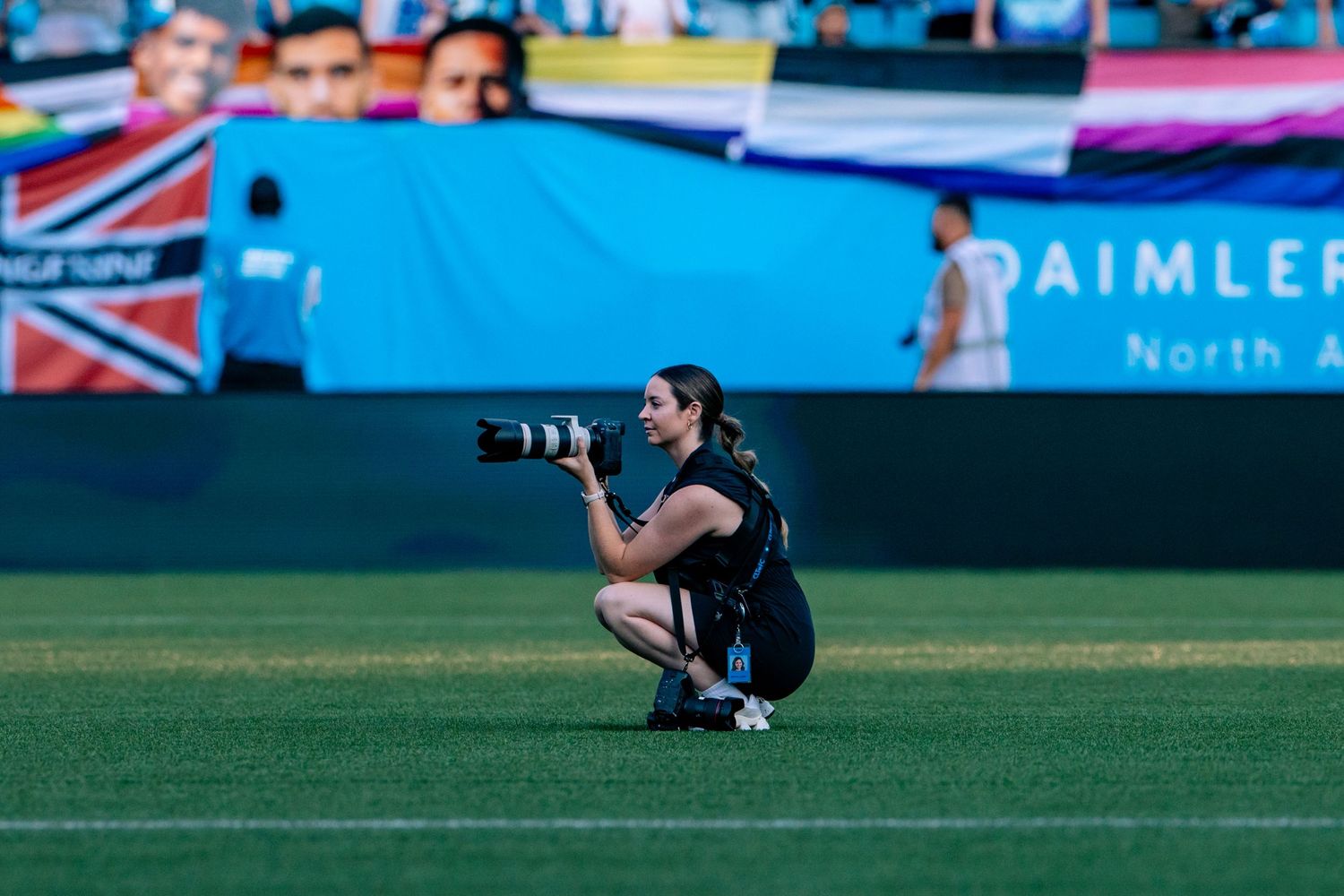 Female sports photographer crouching with professional camera at outdoor soccer stadium.