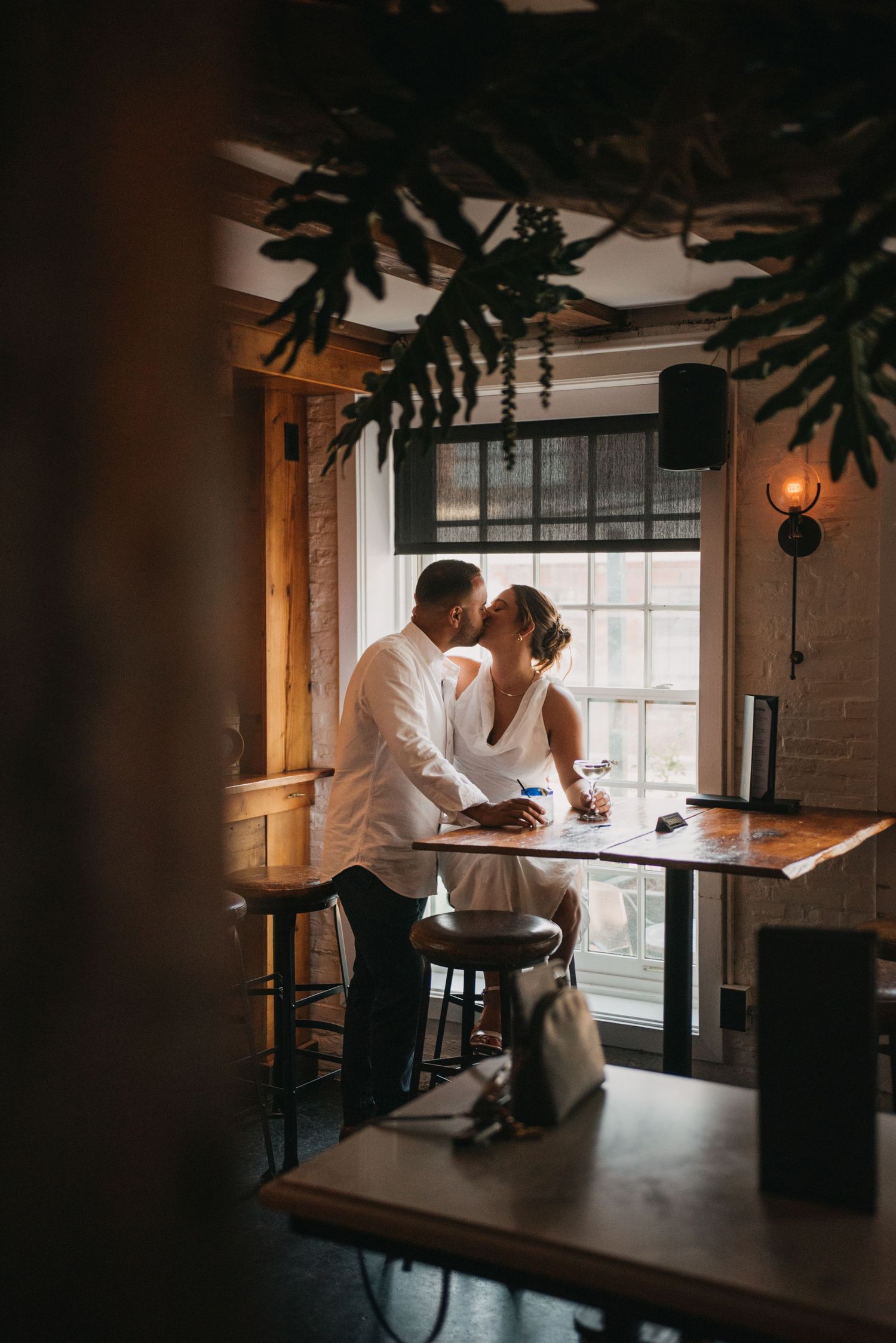 Bride and groom sharing an intimate kiss at a wooden bar table inside a cozy rustic restaurant.
