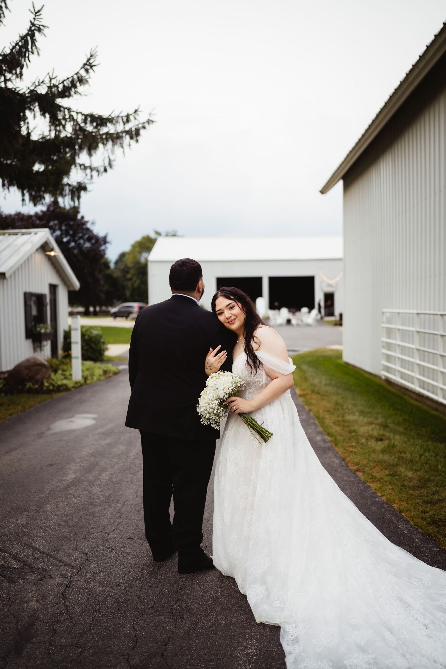 Wedding couple stands on pathway between white barns holding bouquet of white flowers at rustic farm venue.