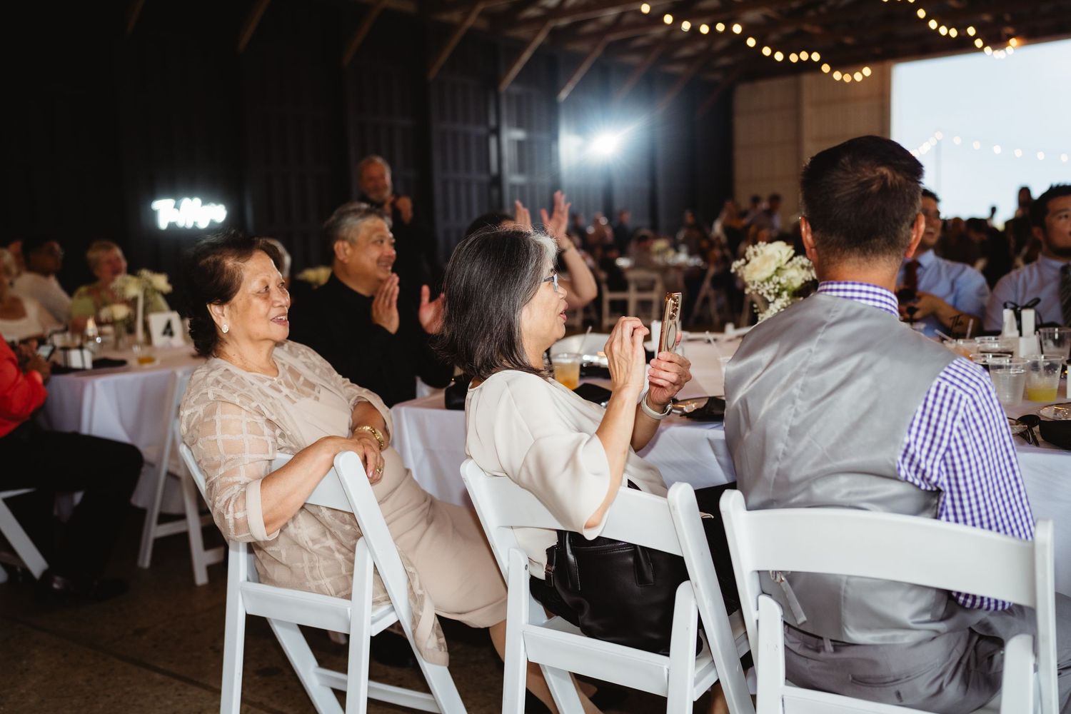 Wedding guests at a reception dining under string lights and white tent decorations at an evening celebration.