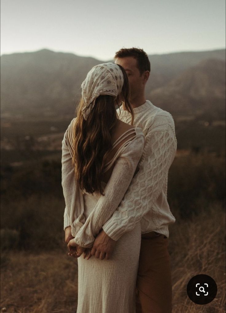 A romantic couple in cozy white sweaters embrace against a misty mountain backdrop at sunset.