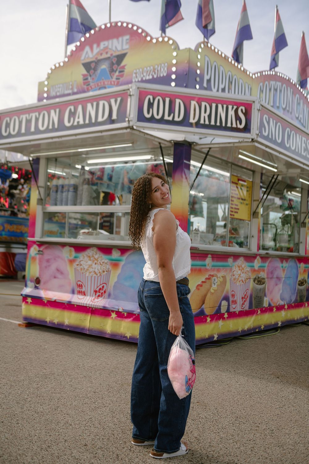 Young woman with curly hair holding cotton candy at a colorful carnival food stand offering cold drinks and sno cones.
