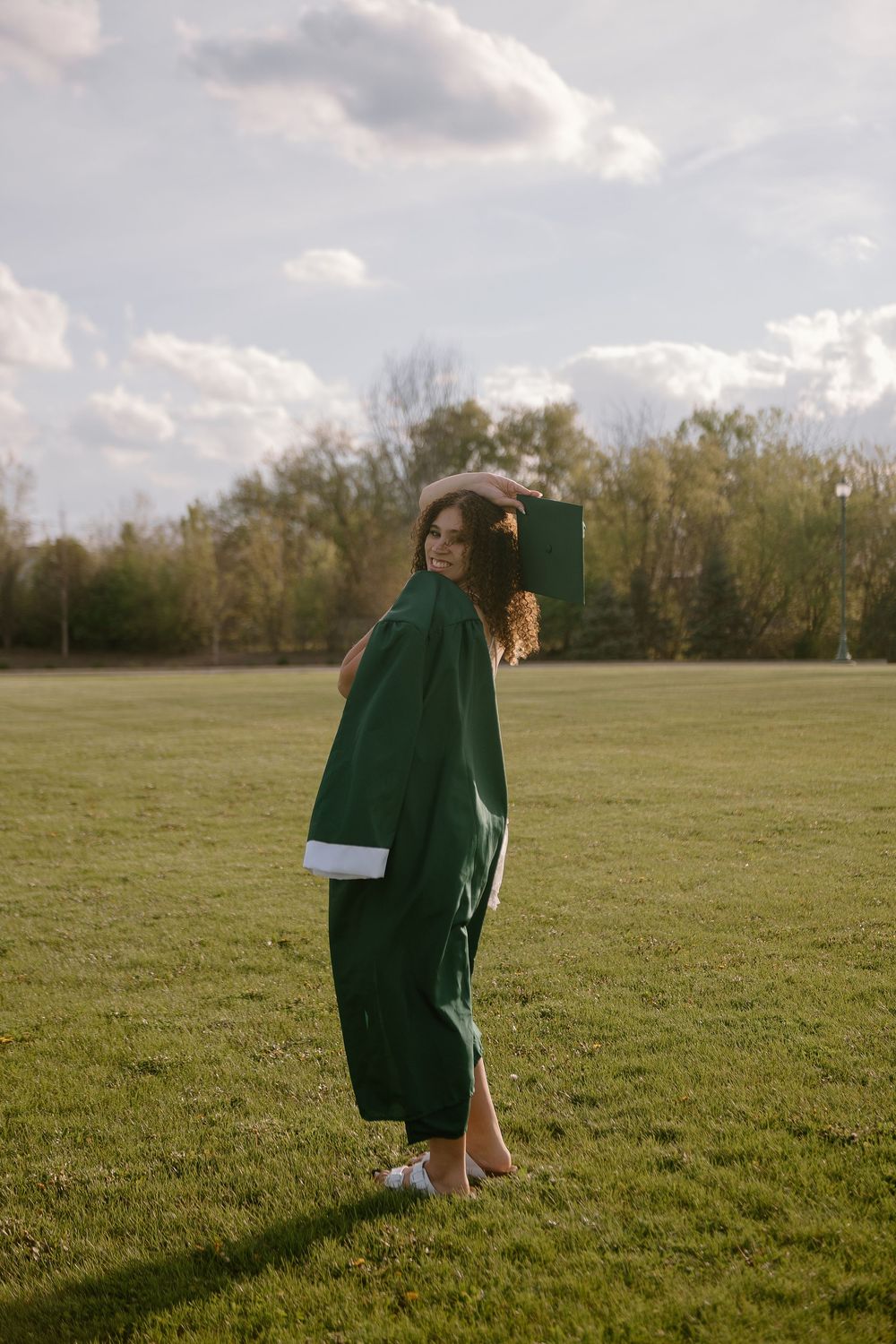 Young woman in green graduation cap and gown standing on grass field with cloudy sky background.