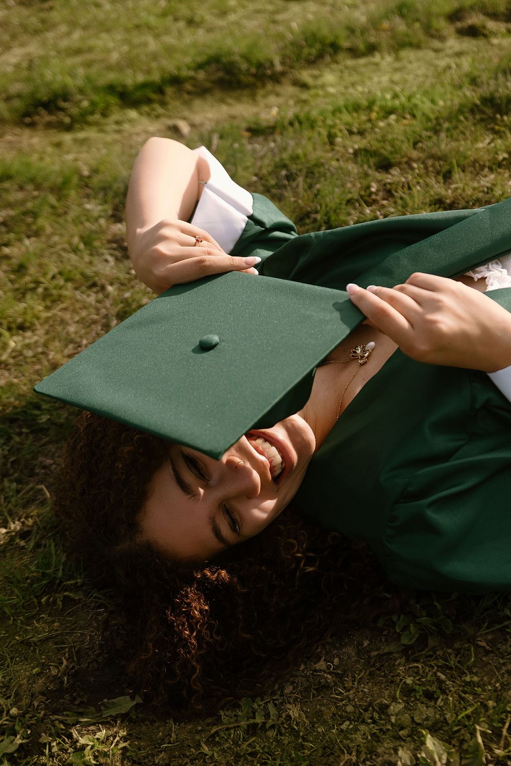 A graduate in green cap and gown lying on grass, smiling while holding her graduation cap over her face.