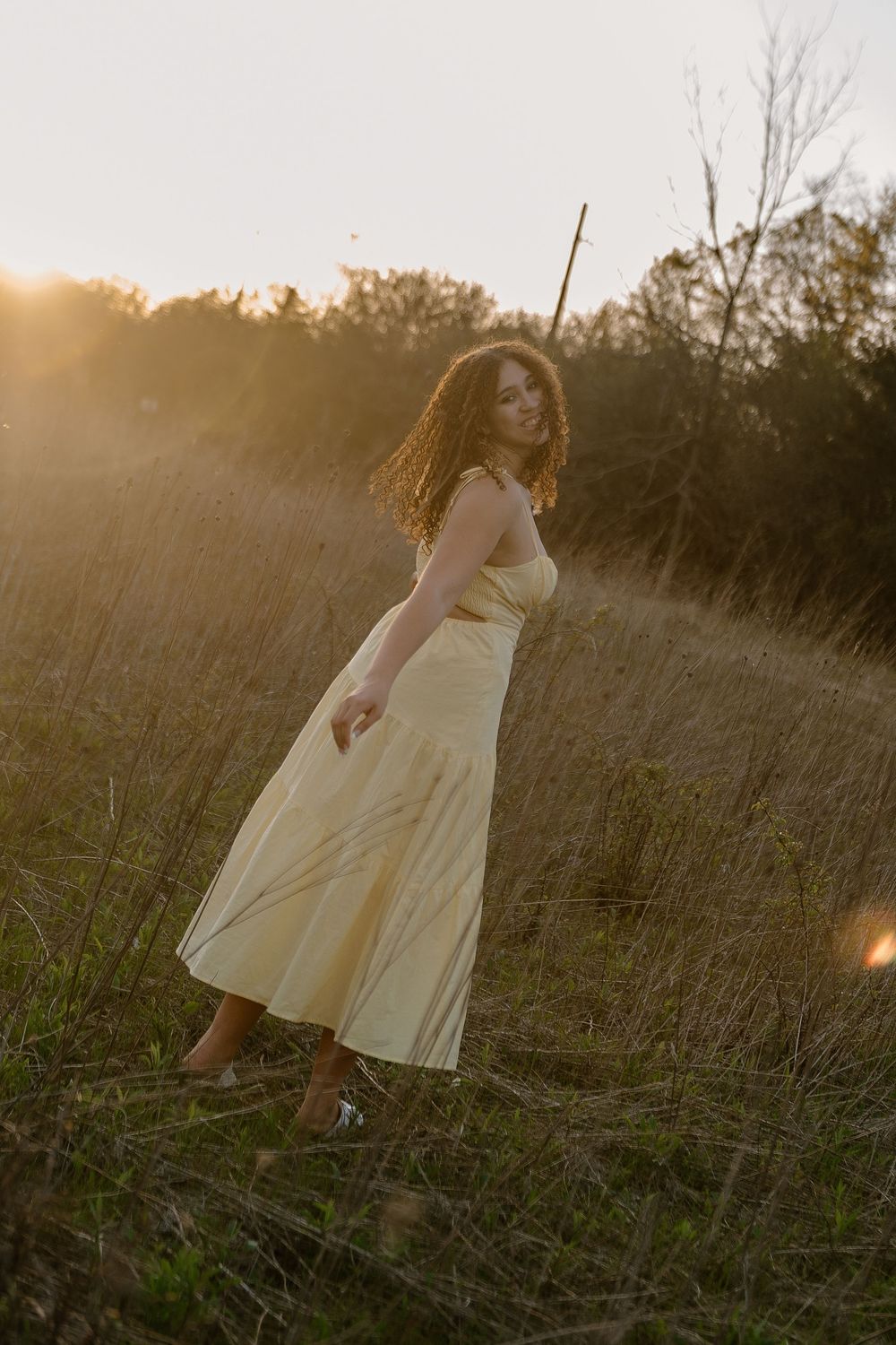 Curly-haired woman in flowing yellow midi dress walks through sunlit meadow at golden hour.