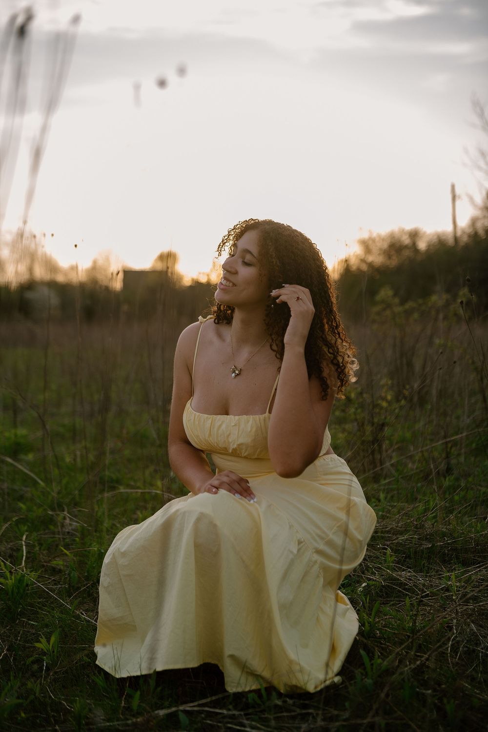 Young woman in yellow dress sitting in field at sunset, curly hair, serene expression, golden hour lighting.