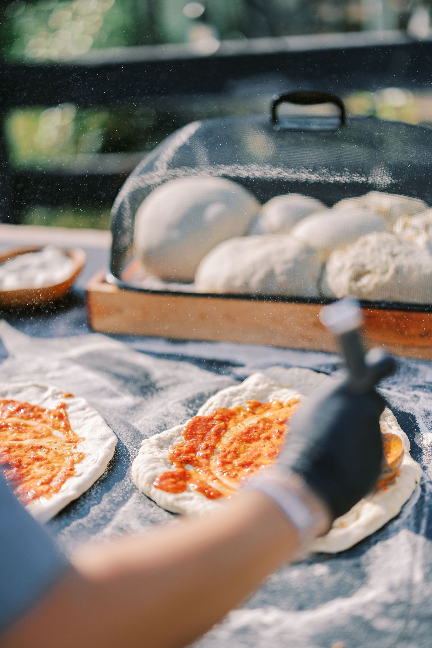 Pizza dough with tomato sauce being prepared on a floured surface with fresh dough under a metal cloche.