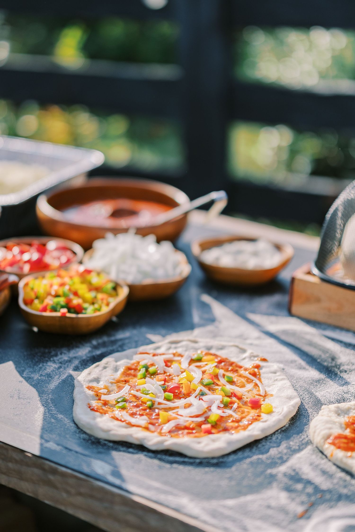 Fresh pizza dough preparation with toppings and ingredients on a blue countertop with various bowls of sauces and veggies.