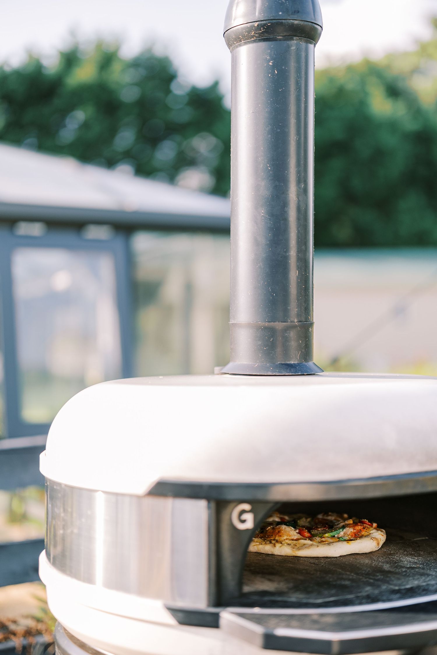 A wood-fired pizza cooking in a sleek outdoor pizza oven with a silver chimney and white dome exterior.