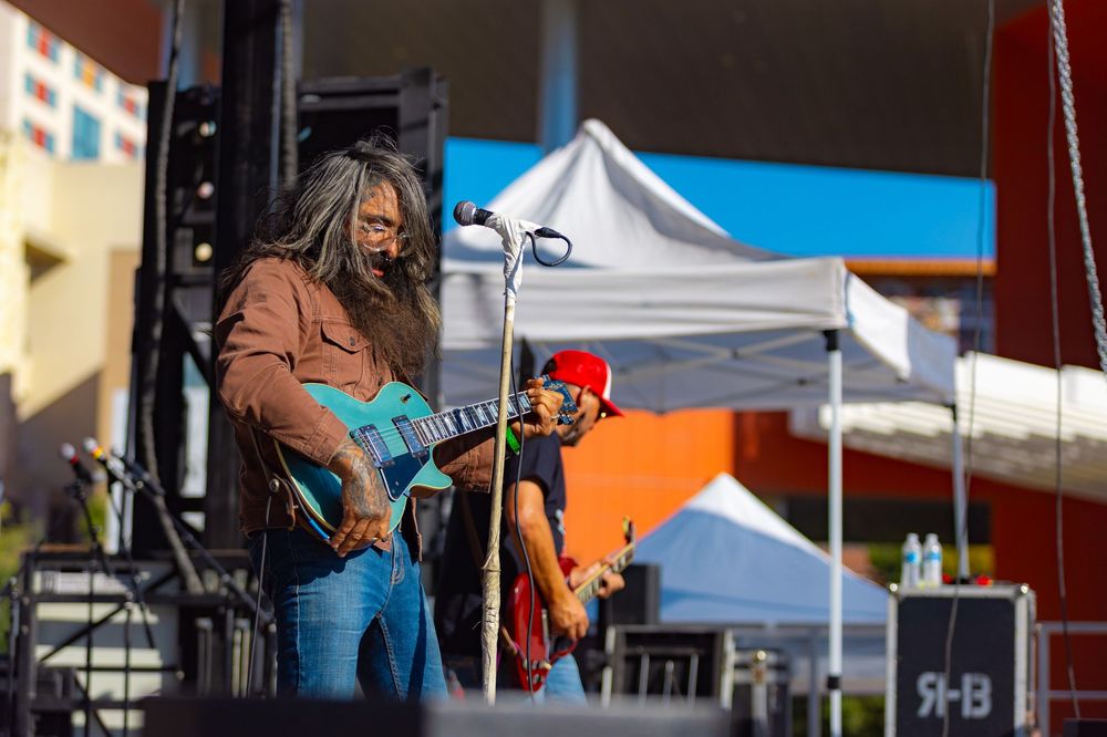Musicians perform on an outdoor stage at a daytime festival with tents and equipment visible in the background.