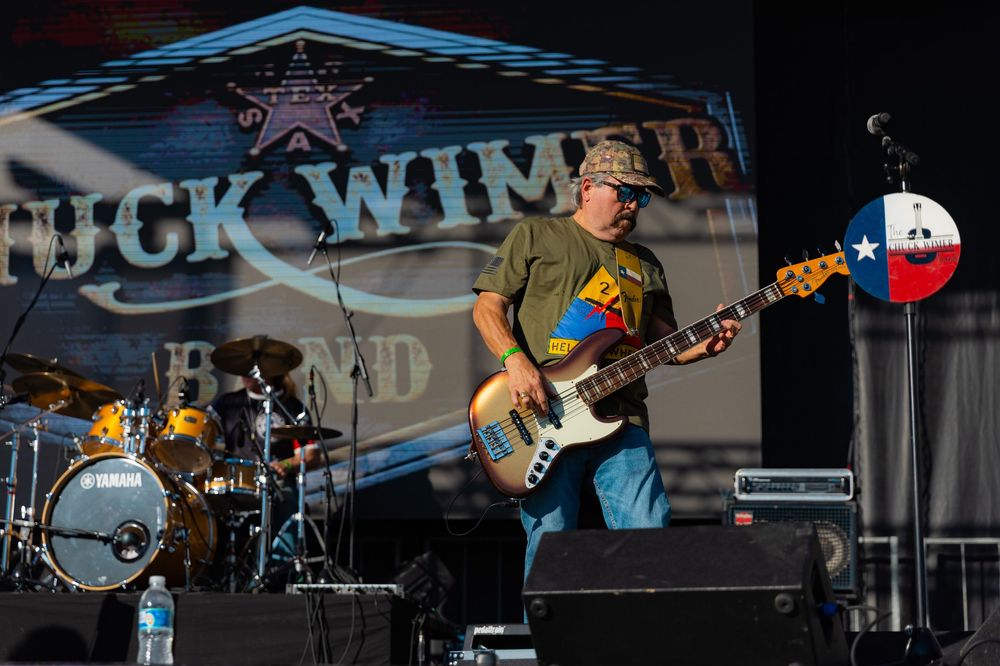 Musicians perform energetically on stage at an outdoor concert with instruments and equipment visible in the background.
