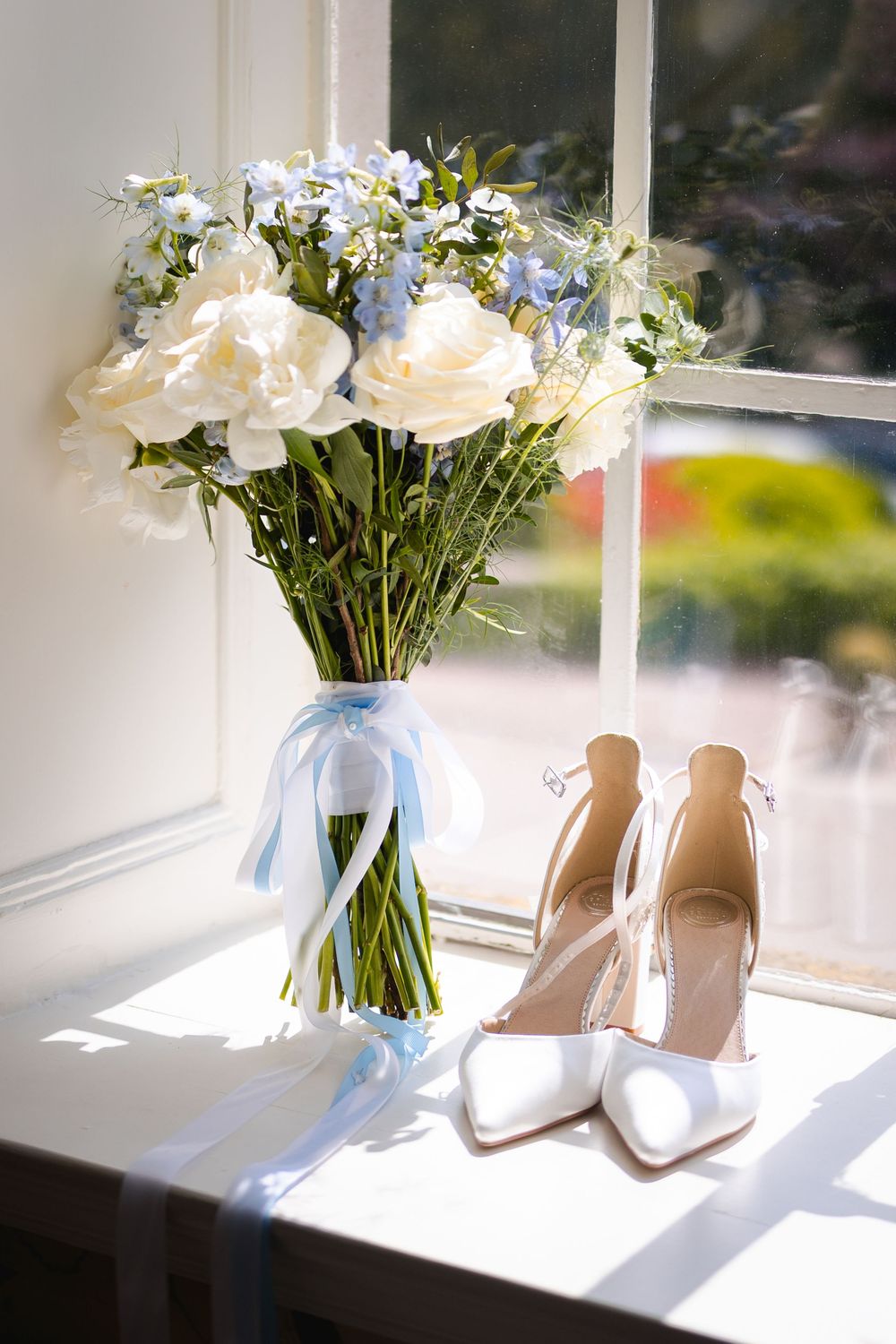 A delicate bridal bouquet with white roses and blue accents sits beside white heels in sunlit window.