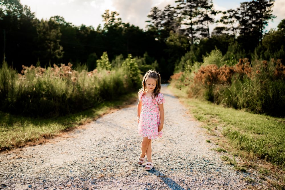A pink-clad figure walks down a gravel path surrounded by lush greenery on a bright summer day.