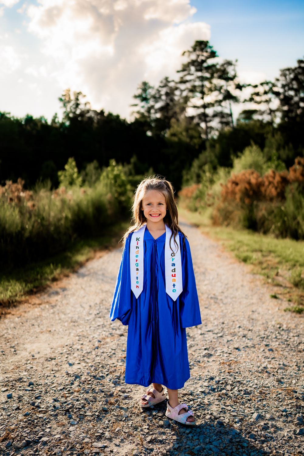 Young graduate in bright blue cap and gown smiles while walking down a gravel path on a sunny day.