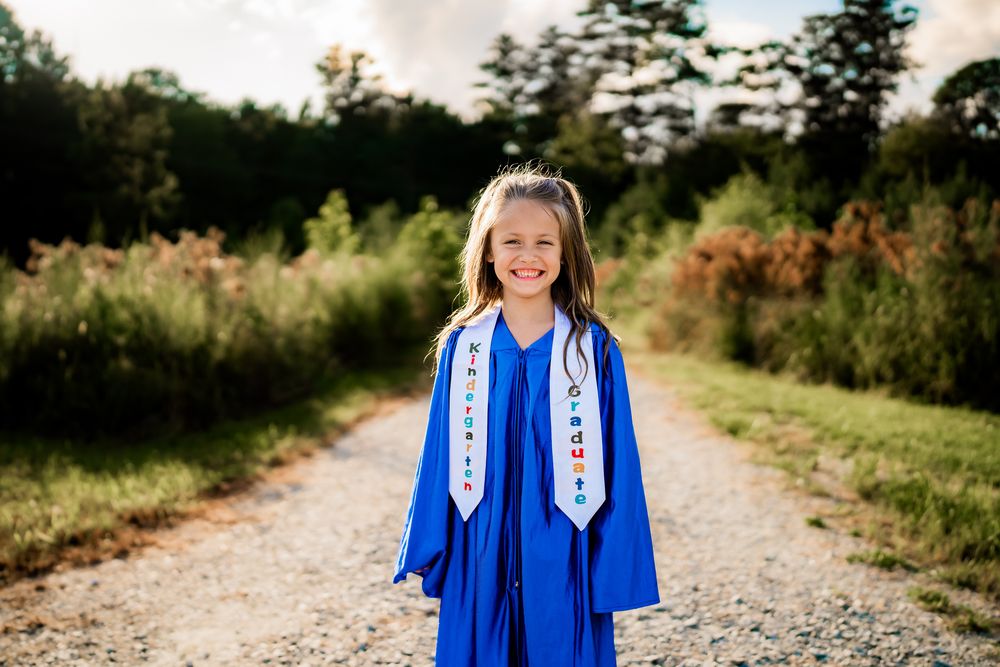 Graduate wearing bright blue cap and gown with white stole stands on gravel path with natural backdrop at sunset.