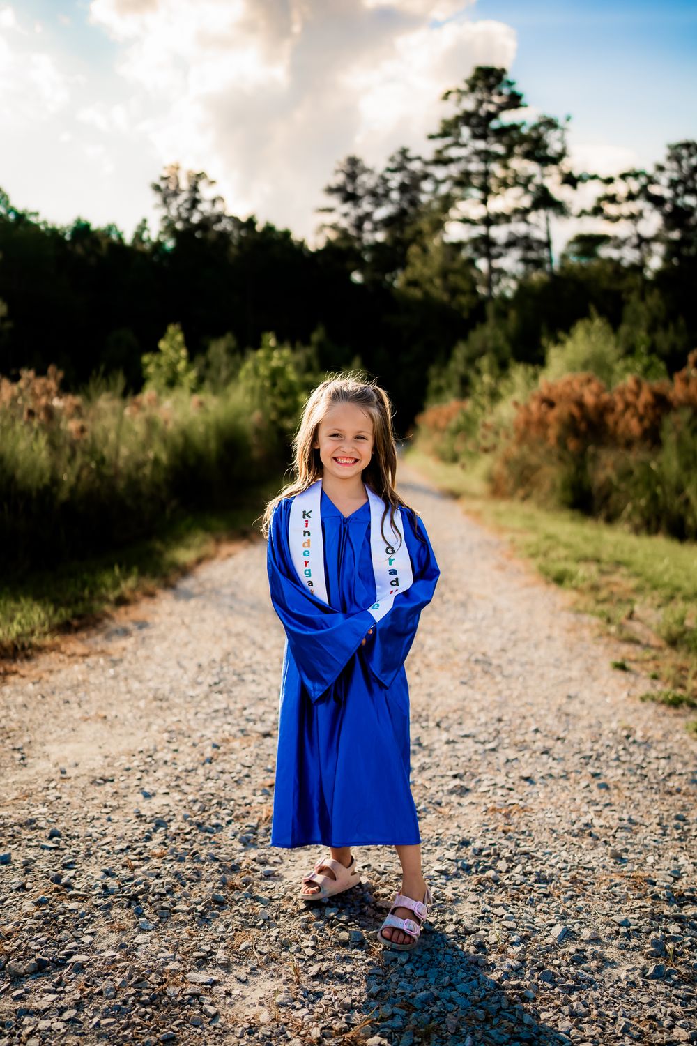 Joyful graduate in bright blue cap and gown poses on a gravel path surrounded by natural scenery at sunset.
