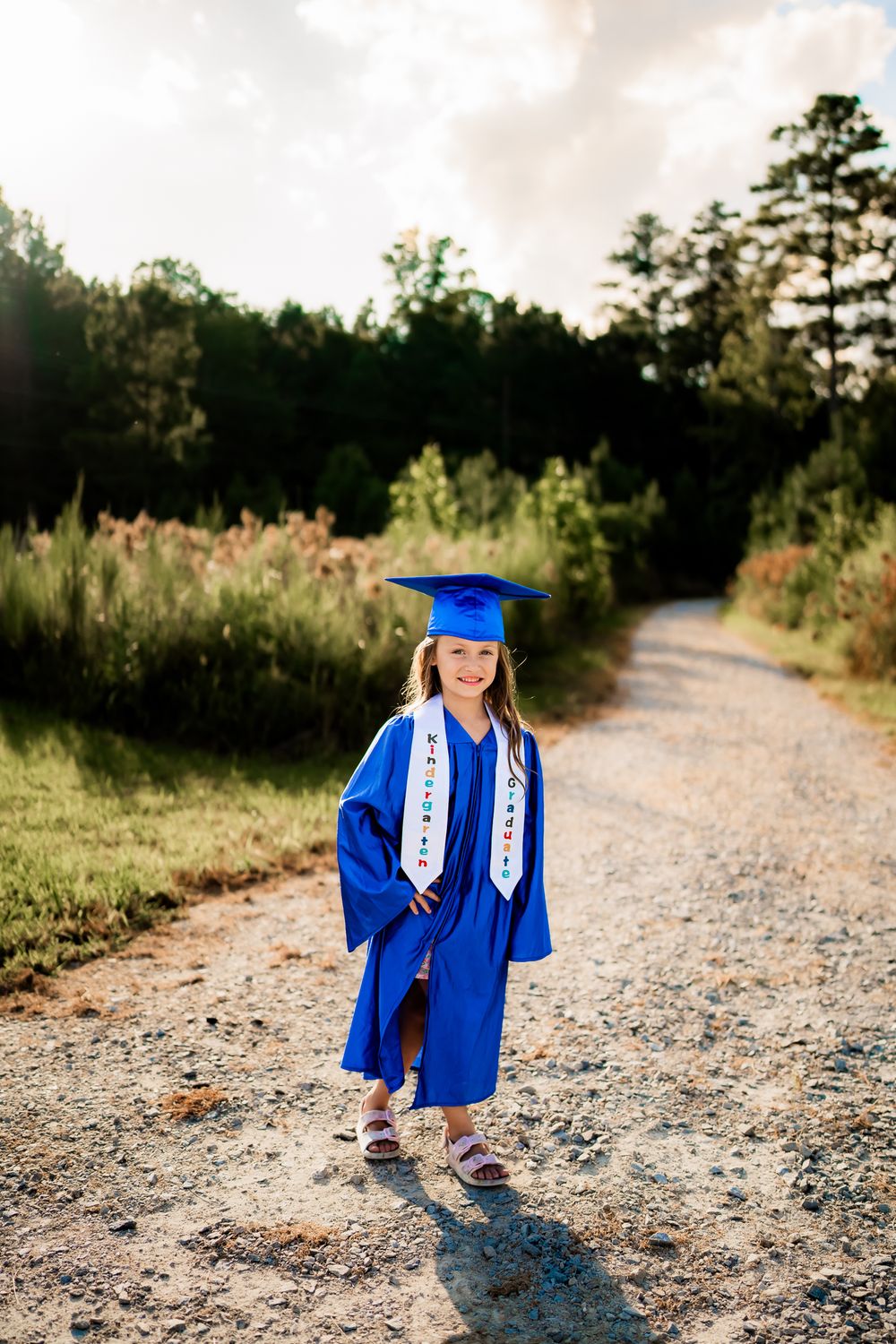 Graduate posing in blue cap and gown on gravel path during outdoor photoshoot session.